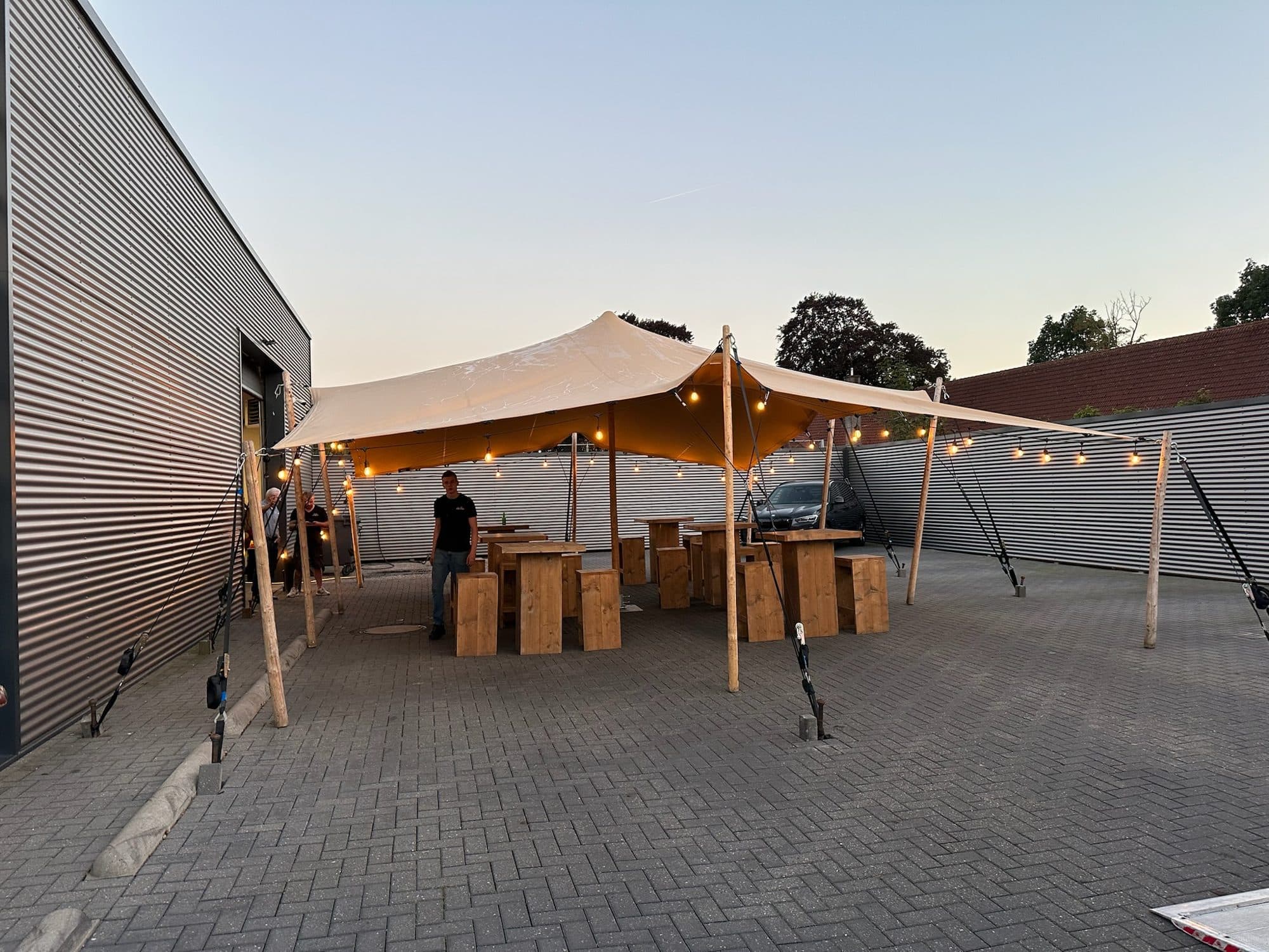 Stretchtent tegen een gevel boven een bestraat terras met houten tafels in de schemering